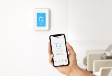 Hand holding a smartphone showing 72 degrees, syncing with a wall-mounted smart thermostat.