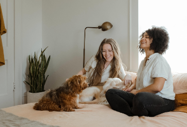 wo women sitting on a bed with their dogs, smiling and enjoying a relaxed moment at home.